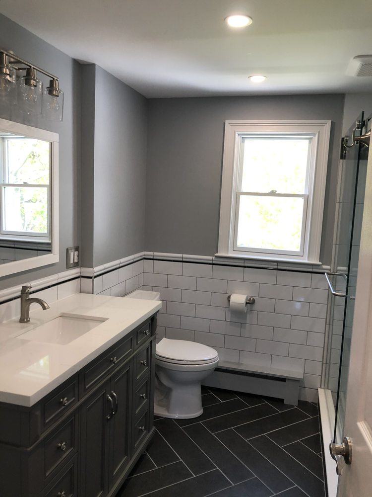 Remodeled bathroom with charcoal herringbone floor tile, white subway tile wainscot, grey vanity and glass shower door