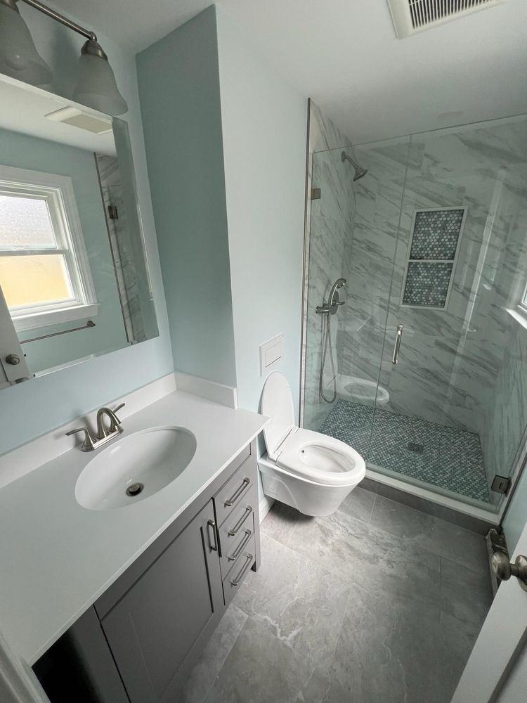 Remodeled bathroom with marble-look porcelain shower walls behind a frameless glass door, a blue-grey mosaic tile niche and matching mosaic shower floor, next to a white vanity and wall-hung toilet