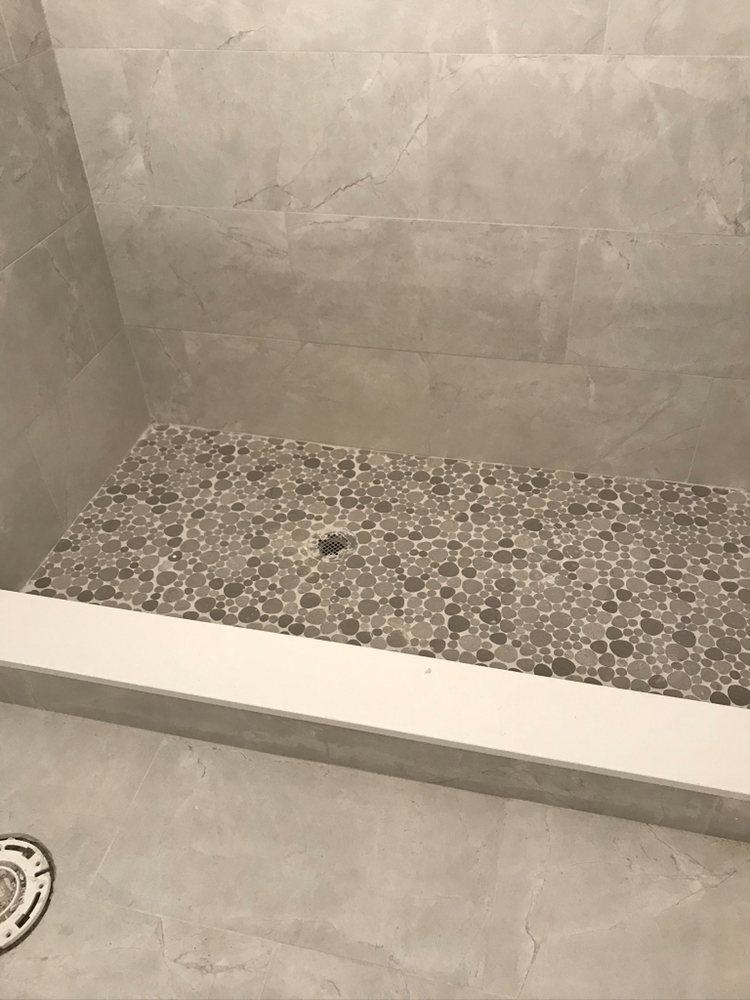 Shower floor of grey pebble mosaic tile with a center drain, framed by large grey porcelain wall tile and a white curb