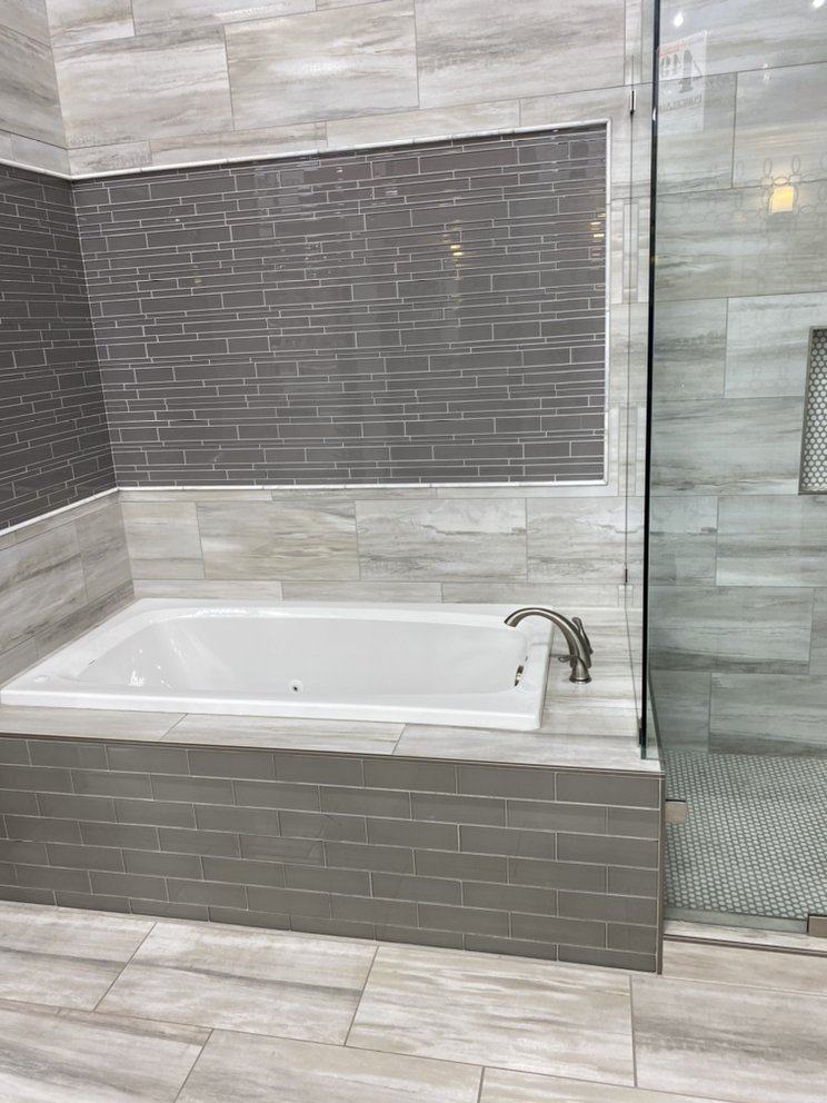 Drop-in tub with a grey linear glass mosaic panel, wood-look porcelain wall and floor tile, and tiled tub apron